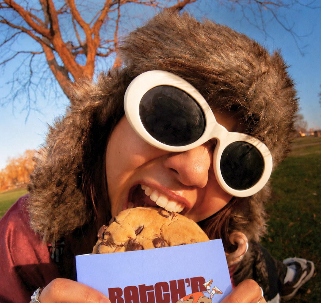 A person outdoors wearing a brown fur-trimmed winter hat and white oval sunglasses, holding a large chocolate chip cookie toward their mouth while holding a blue card with the word 'BATCH' in red letters.